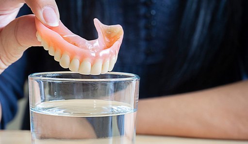 Fingers placing upper arch of dentures into glass of clear fluid